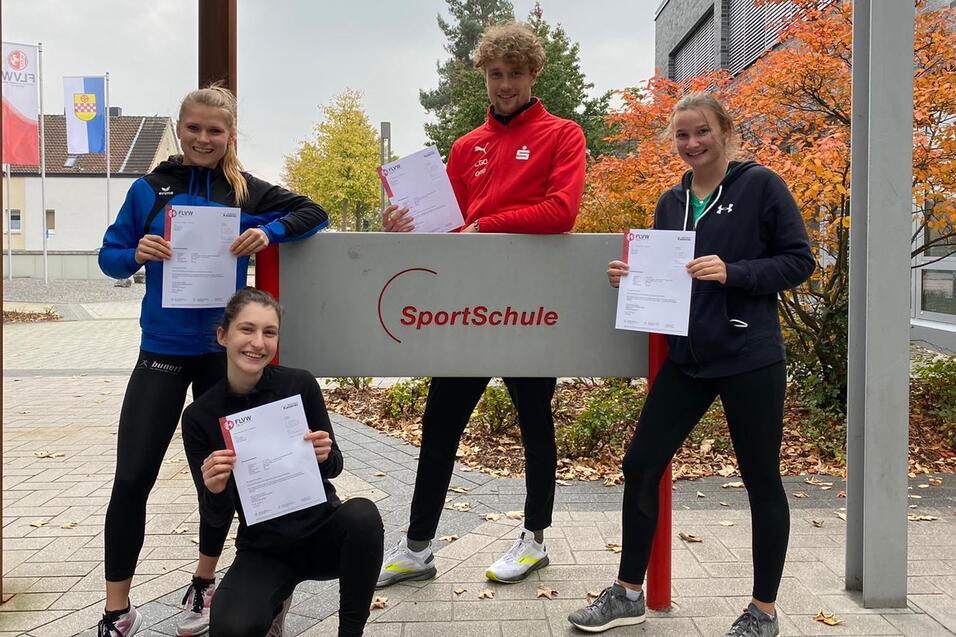 Vier junge Erwachsene halten Zertifikate vor dem Eingang einer SportSchule, umgeben von B&auml;umen und Geb&auml;uden.