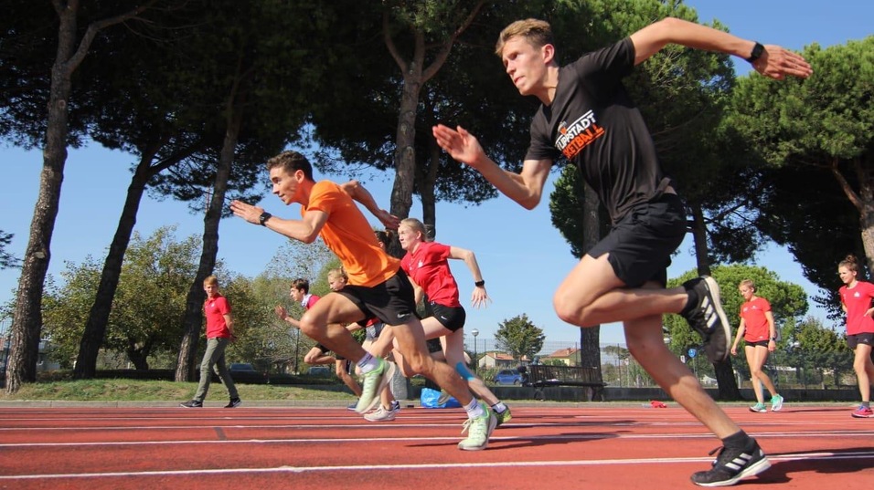 Sechs Athleten sprinten auf einer roten Laufbahn unter blauem Himmel und begr&uuml;nten B&auml;umen.
