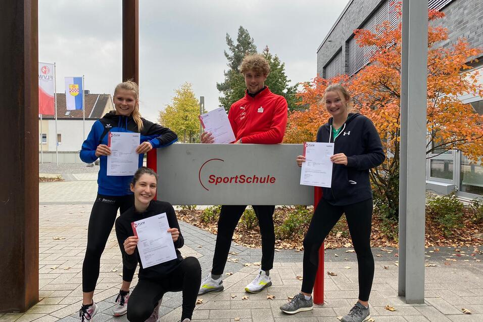 Vier junge Erwachsene halten Zertifikate vor dem Eingang einer SportSchule, umgeben von B&auml;umen und Geb&auml;uden.