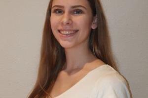 Junge Frau mit langen, braunen Haaren, l&auml;chelt vor grauer Wand. Sie tr&auml;gt ein helles, schlichtes Oberteil.