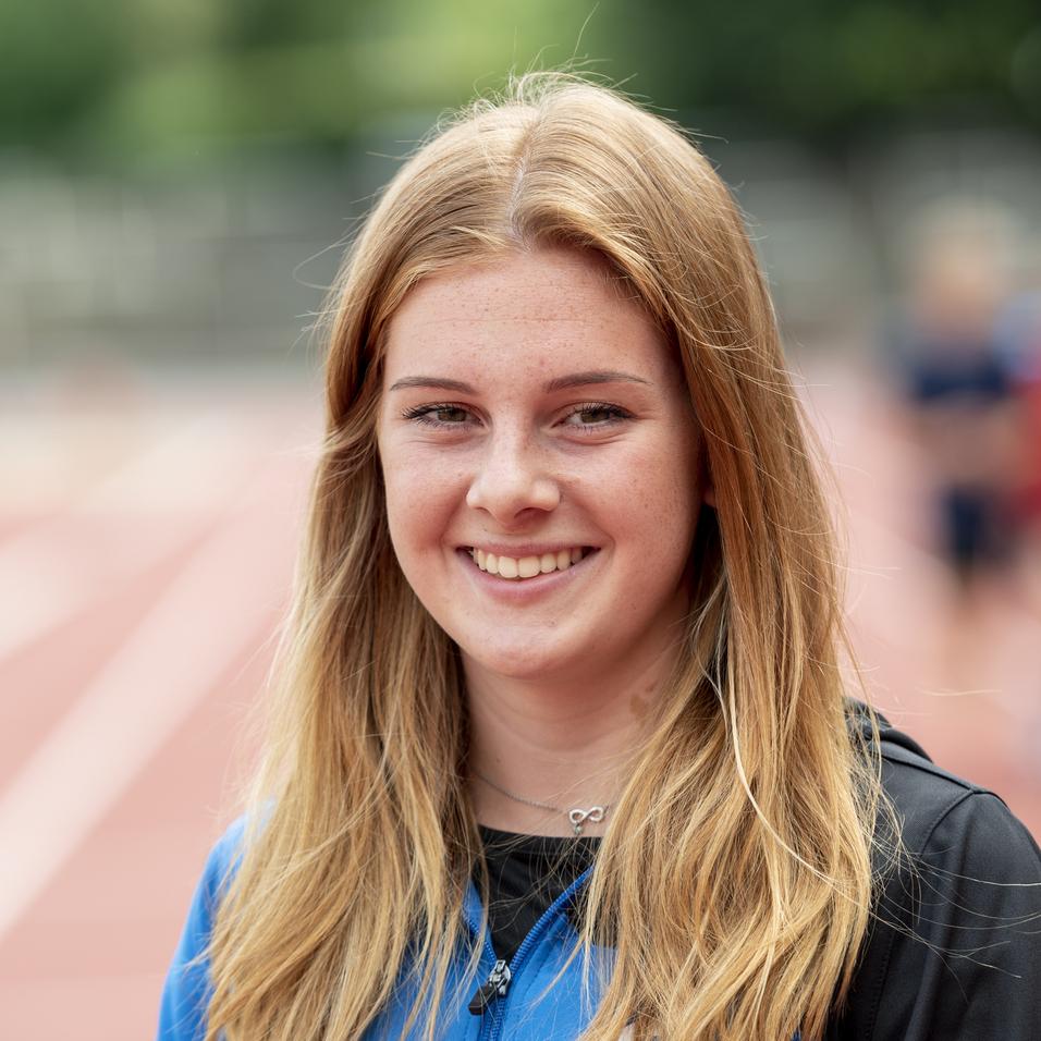 Lächelnde junge Frau in blauer Sportjacke steht auf einer Laufbahn im Freien, unscharfer Hintergrund mit Sportlern.