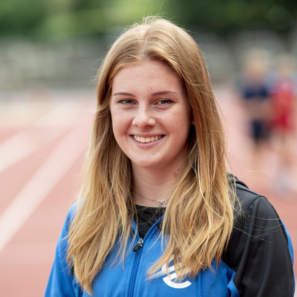 Lächelnde junge Frau in blauer Sportjacke steht auf einer Laufbahn im Freien, unscharfer Hintergrund mit Sportlern.