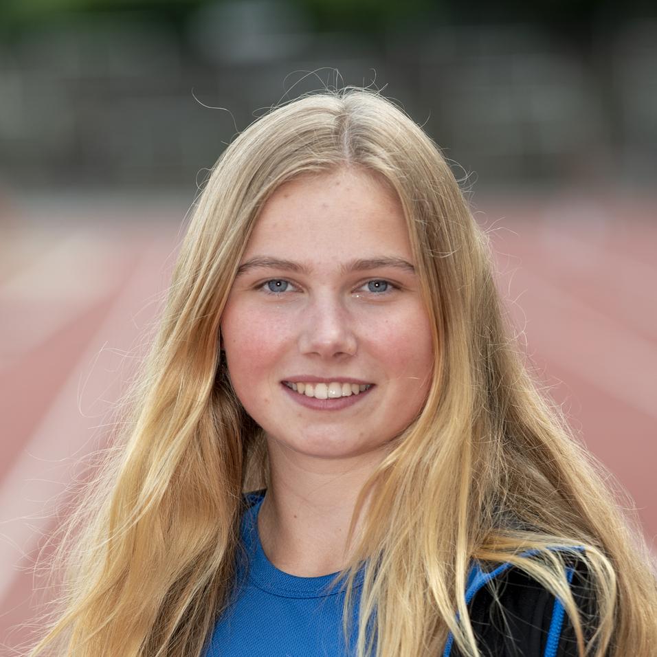 Junge Sportlerin mit langen blonden Haaren steht auf einer roten Laufbahn, trägt ein blaues Sportoberteil und eine Jacke.