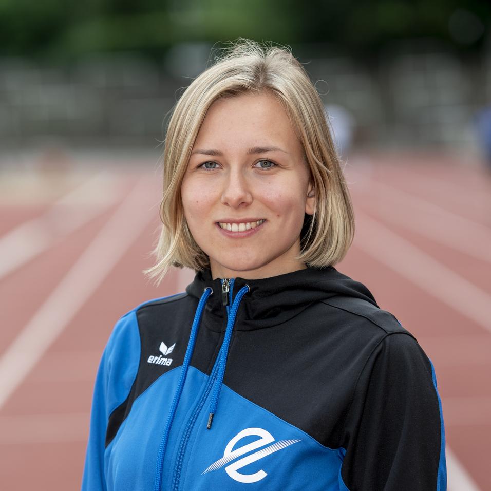 Junge Athletin in blau-schwarzer Sportjacke steht auf einer Laufbahn. Unscharfer Hintergrund mit weiteren Sportlern.