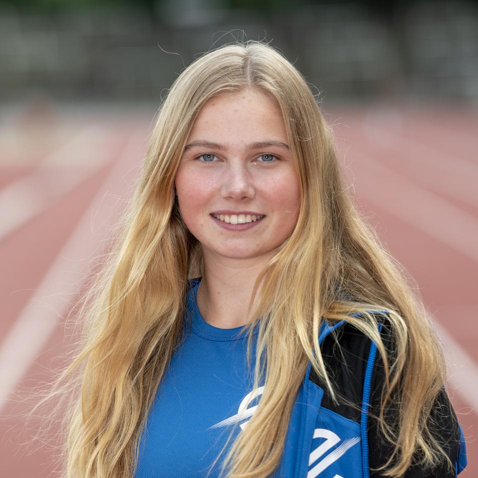 Junge Sportlerin mit langen blonden Haaren steht auf einer roten Laufbahn, trägt ein blaues Sportoberteil und eine Jacke.