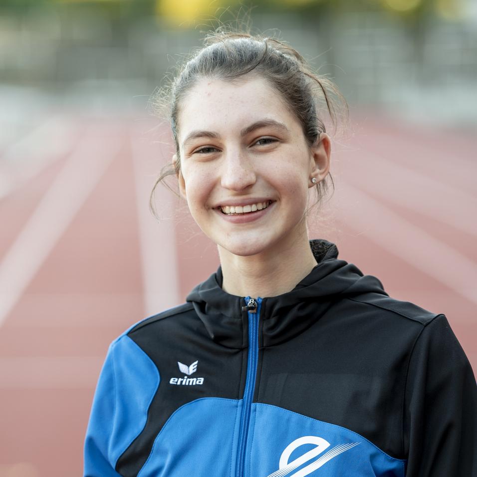 Lächelnde Athletin mit Sportjacke steht auf einer Laufbahn im Freien, verschwommener Hintergrund zeigt weitere Sporteinrichtungen.