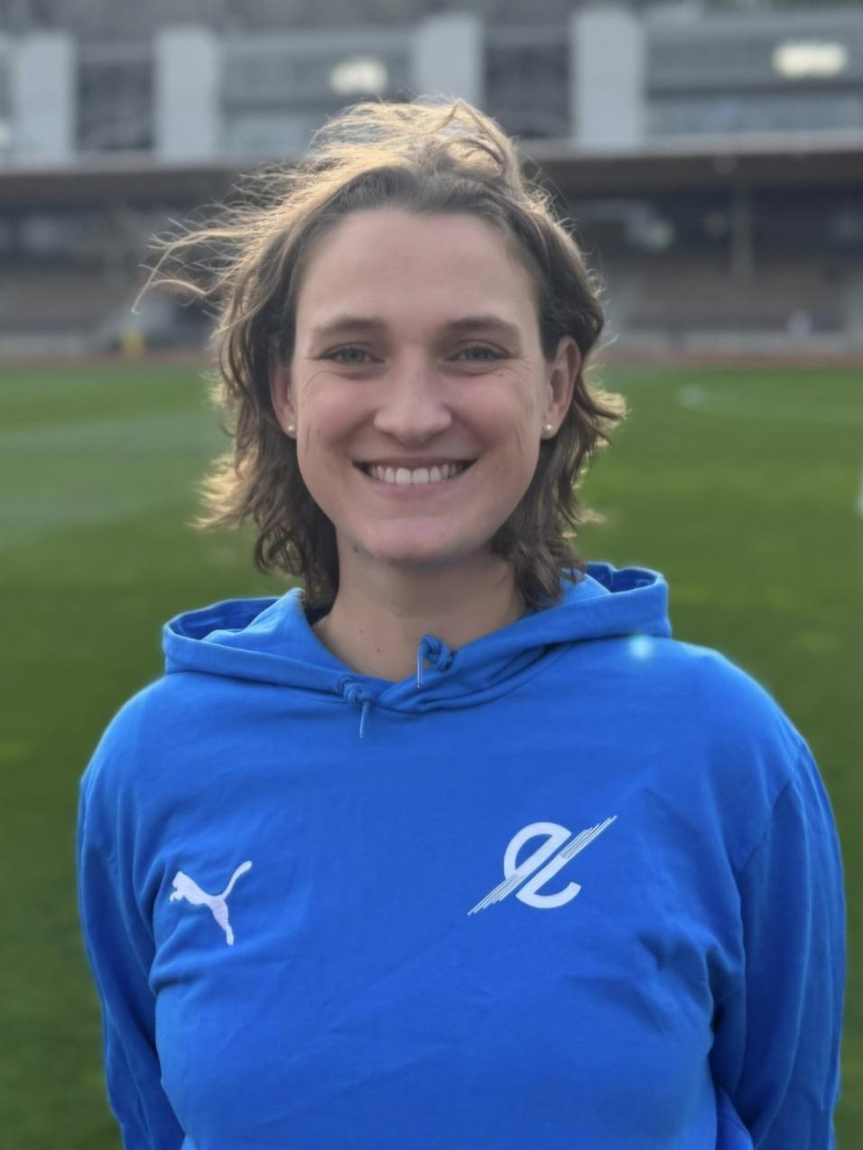 Lächelnde Person mit kurzen Haaren trägt einen blauen Hoodie auf einem Sportfeld im Hintergrund.