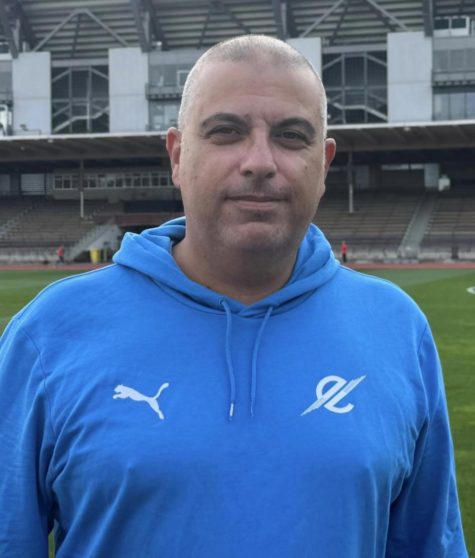 Porträt eines Mannes in blauer Kapuzenjacke, stehend auf einem Sportfeld mit Stadion im Hintergrund.