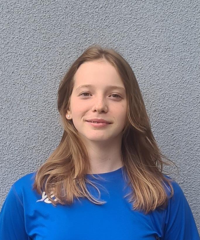 Porträt einer jungen Frau mit langen Haaren, die ein blaues Sportshirt trägt, vor einer grauen Wand.
