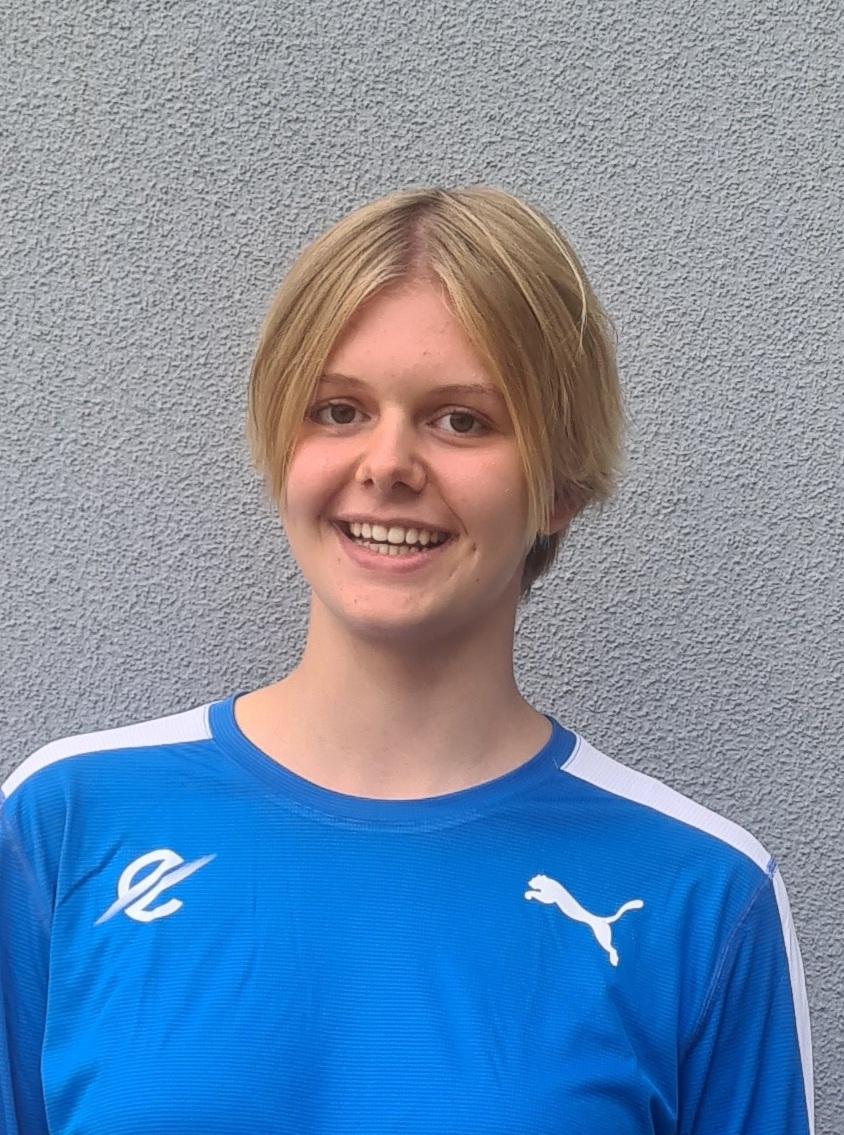 Junge Frau mit kurzer blonder Frisur trägt ein blaues Sportshirt vor grauem Hintergrund.
