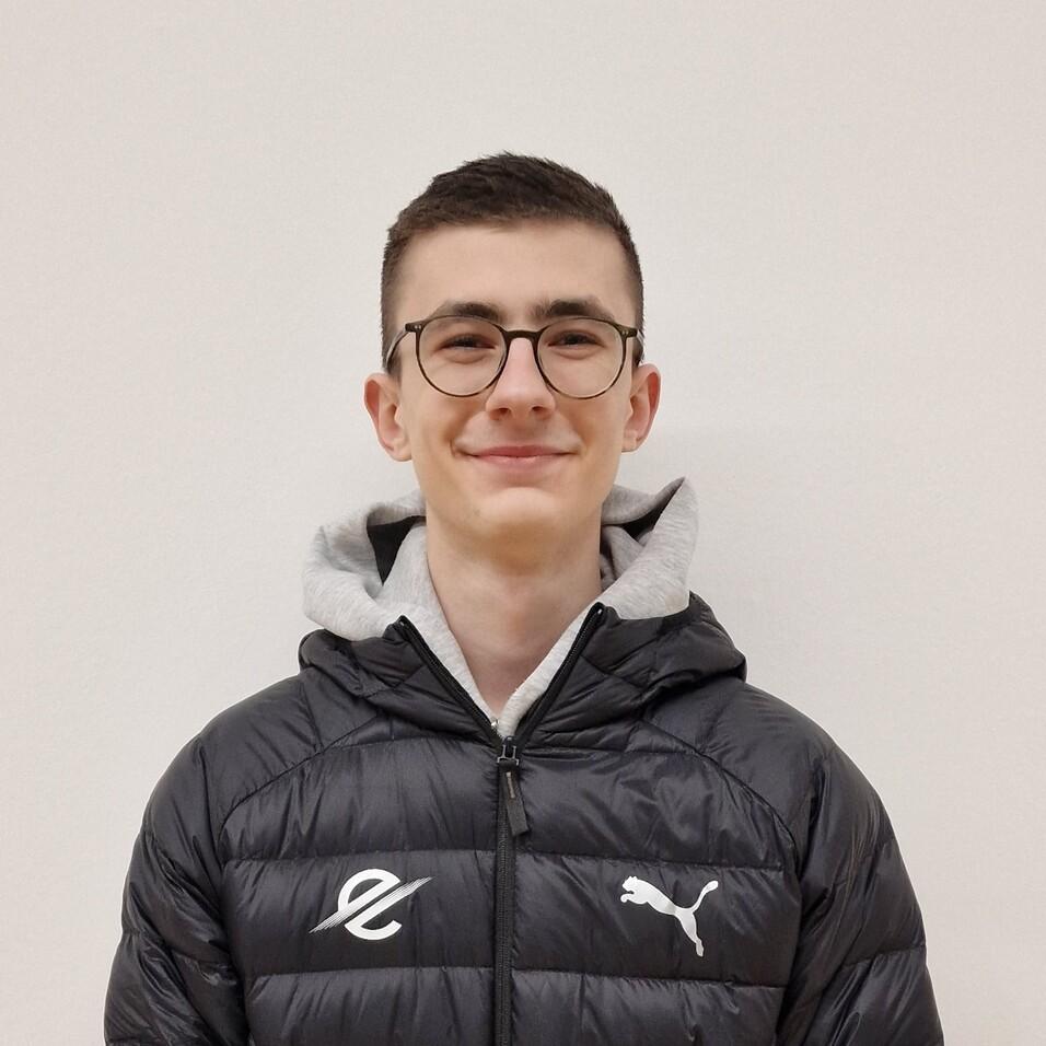 Junger Mann mit Brille tr&auml;gt eine schwarze, gepolsterte Jacke und steht vor einer hellen Wand.