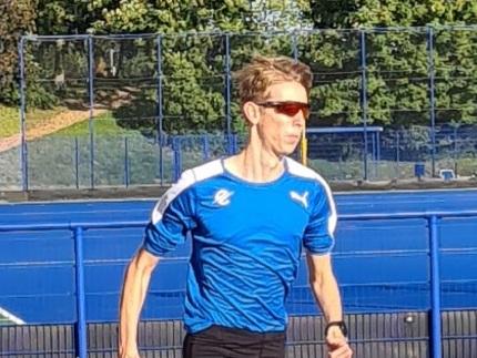 Sportler in blauer Laufbekleidung joggt auf einer Laufbahn, im Hintergrund B&auml;ume und Sporteinrichtungen.