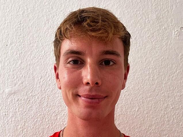 Junger Mann mit blonden, lockigen Haaren, tr&auml;gt ein rotes Shirt, steht vor einer wei&szlig;en Wand.
