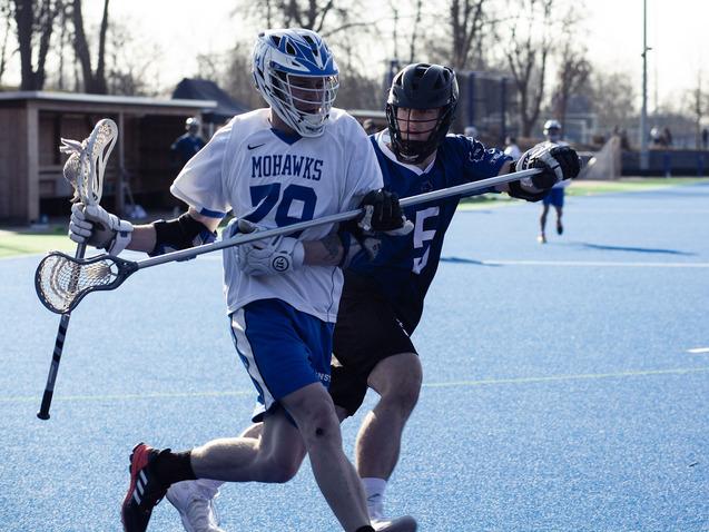 Lacrosse-Spieler der Mohawks dribbelt mit Ball, w&auml;hrend ein Gegner ihn verfolgt. Beide tragen Schutzhelme und Sportkleidung.
