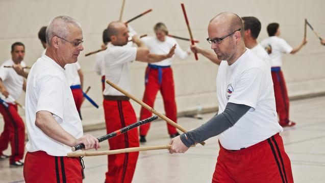 Zwei Männer in Trainingsanzügen üben mit langen Stöcken in einer Sporthalle, umgeben von weiteren Teilnehmern.