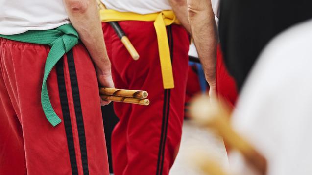 Nahaufnahme von Personen in roten Hosen mit farbigen Gürteln, die Holzstöcke in den Händen halten.