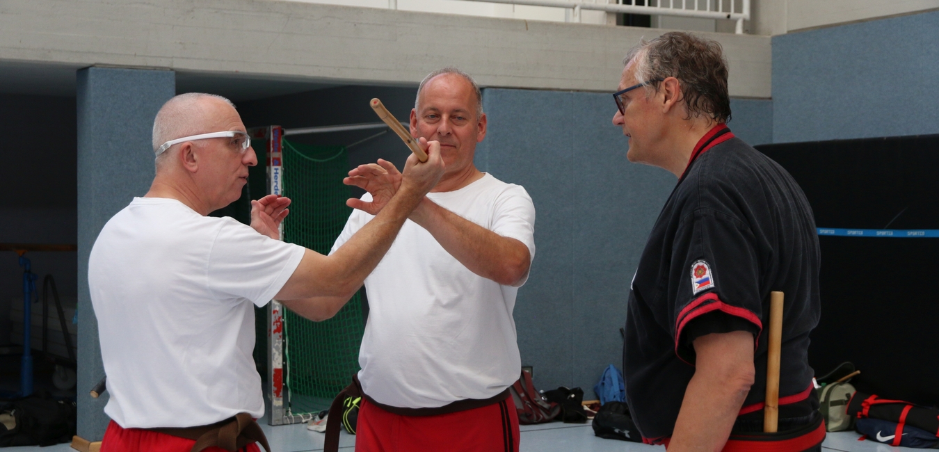 Drei M&auml;nner in Trainingsanz&uuml;gen &uuml;ben mit St&ouml;cken in einer Sporthalle.