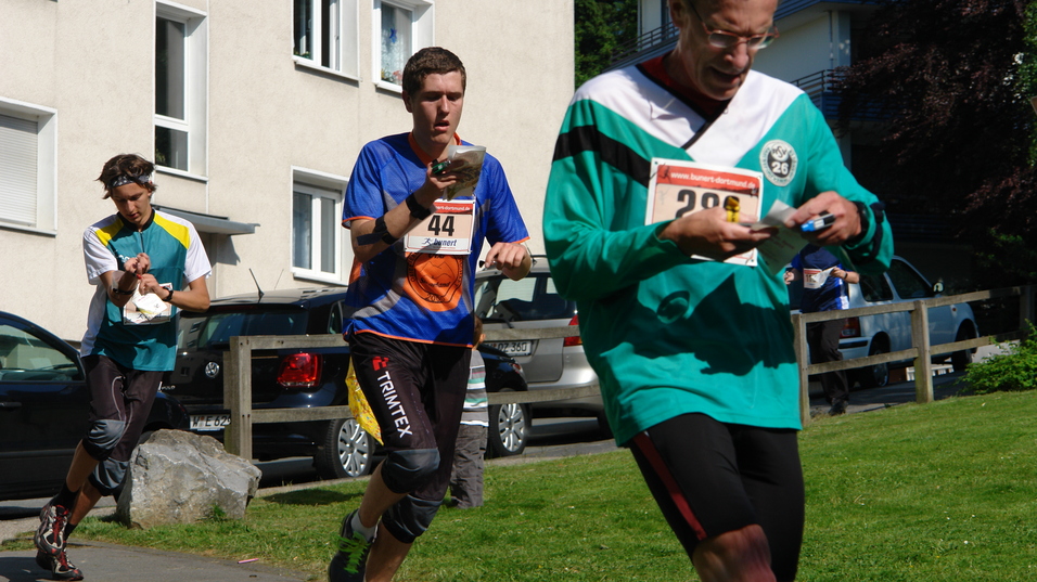 Drei M&auml;nner laufen auf einem Weg, zwei tragen Startnummern und eine sportliche Kleidung. Wohngeb&auml;ude im Hintergrund.