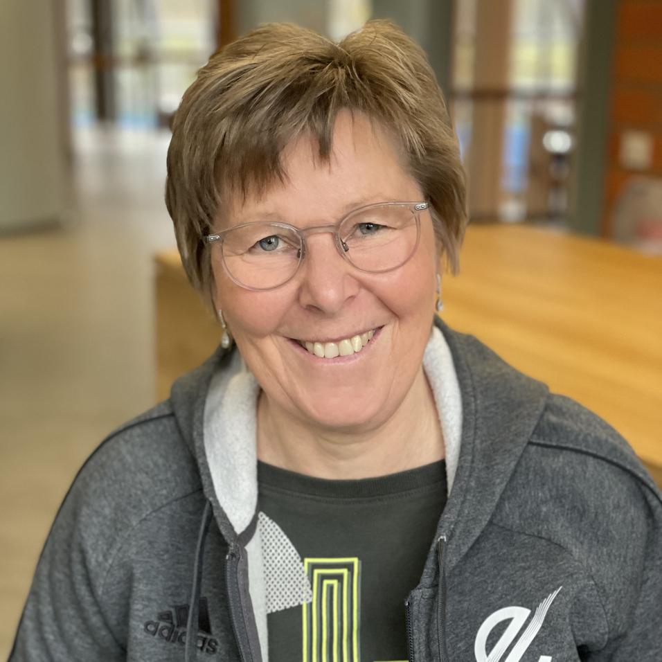 Lächelnde Frau mit kurzen Haaren in grauer Jacke und Brille sitzt in einem modernen Innenraum. Hintergrund unscharf.