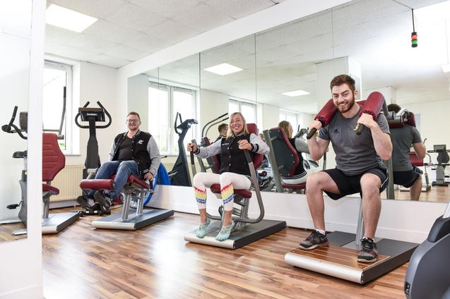 Drei Personen trainieren auf Fitnessger&auml;ten in einem modernen Gym mit gro&szlig;en Spiegeln und hellen W&auml;nden.