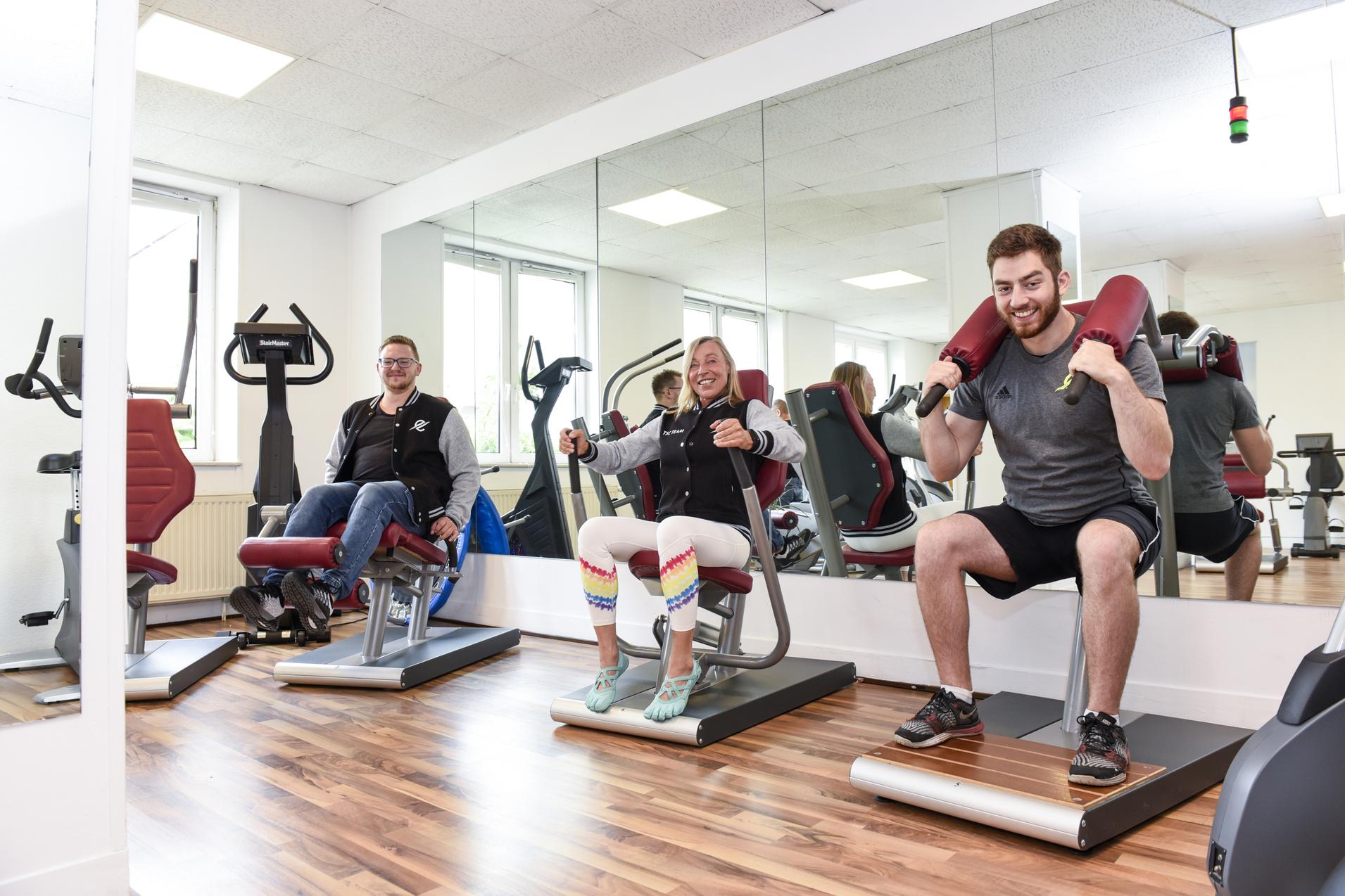 Drei Personen trainieren auf Fitnessger&auml;ten in einem modernen Gym mit gro&szlig;en Spiegeln und hellen W&auml;nden.