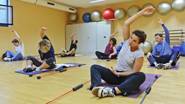 Gruppe von sechs Personen bei einem Fitness-Training mit Gymnastikbällen und Übungsgeräten in einem hellen Raum.
