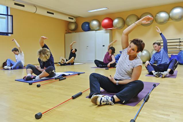Gruppe von sechs Personen bei einem Fitness-Training mit Gymnastikbällen und Übungsgeräten in einem hellen Raum.