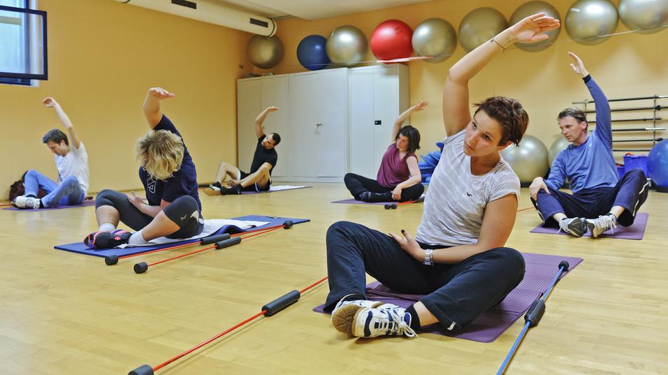Gruppe von sechs Personen bei einem Fitness-Training mit Gymnastikbällen und Übungsgeräten in einem hellen Raum.