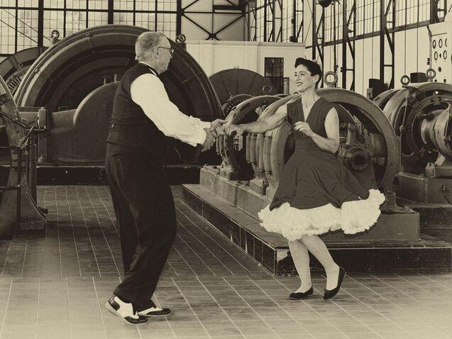 Tanzendes Paar in vintage Kleidung vor großen Maschinen in einem Industriegebäude.