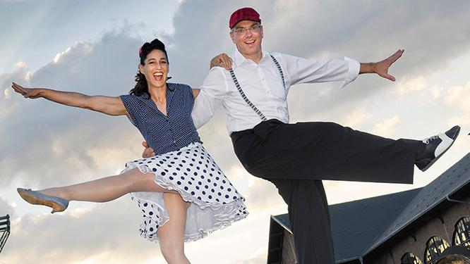 Tanzendes Paar in fr&ouml;hlicher Pose vor einem bew&ouml;lkten Himmel, ein Mann und eine Frau im Retro-Stil mit H&uuml;ten.