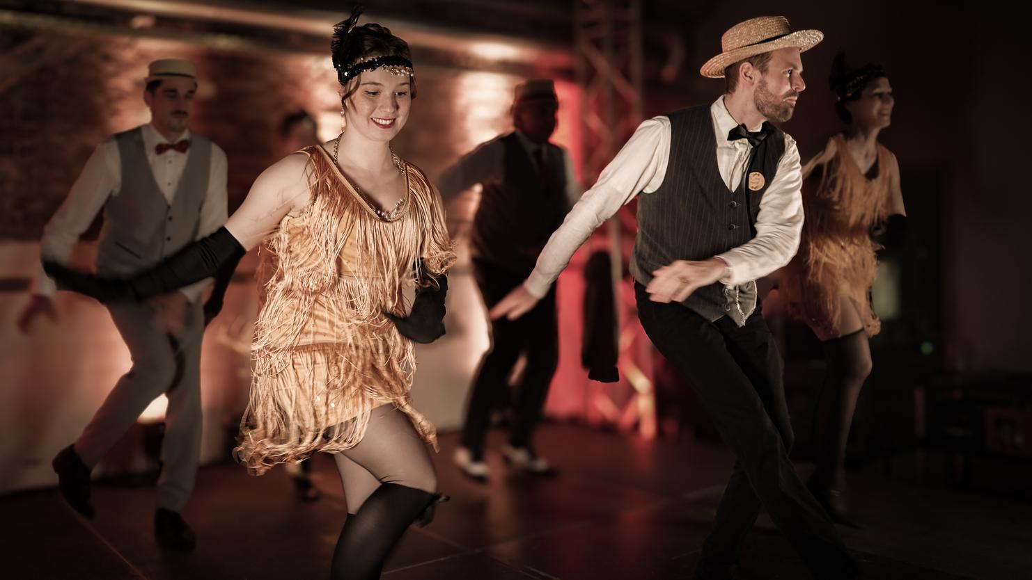 Tanzende Personen im Vintage-Stil mit Fransenkleidung und Hüten auf einer Bühne im gedimmten Licht.