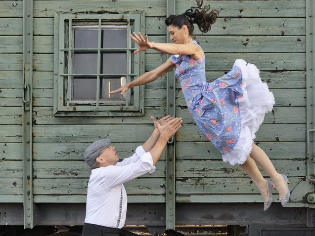 Tanzpaar in vintage Kleidung: Frau springt zu Mann vor einer grünen Eisenbahnwagenwand.