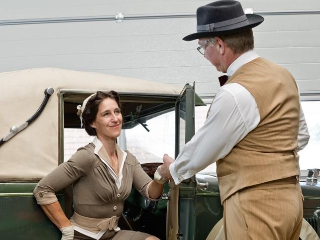Frau in vintage Outfit steigt elegant aus einem Oldtimer, während ein Mann im Anzug ihr die Hand reicht.