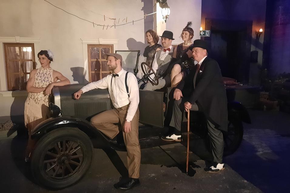 Sechs Personen in vintage Kleidung posieren um ein altes Auto vor einer historischen Kulisse bei ged&auml;mpftem Licht.
