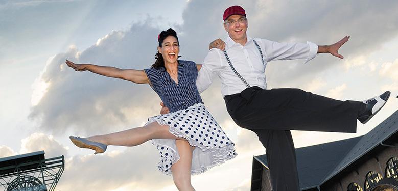 Tanzendes Paar in fröhlicher Pose vor einem bewölkten Himmel, ein Mann und eine Frau im Retro-Stil mit Hüten.