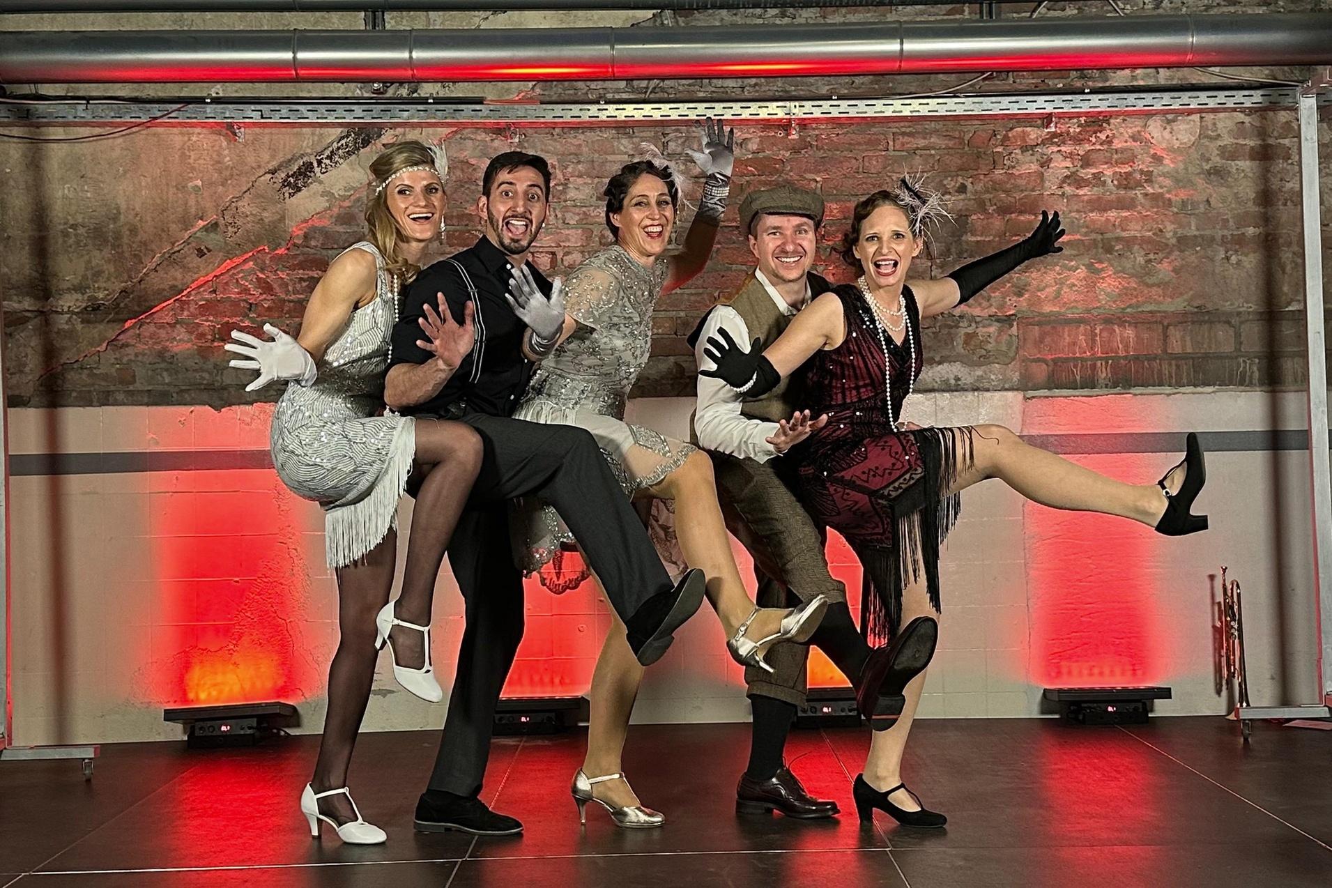 F&uuml;nf Personen in vintage Tanzkleidung posieren auf einer B&uuml;hne und heben die Beine in einer dynamischen Tanzbewegung.