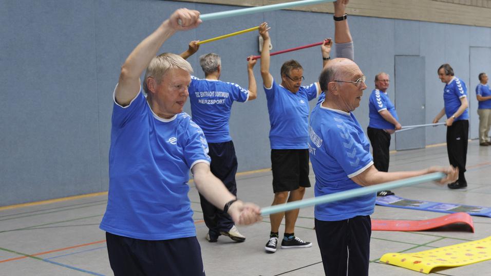 Ältere Männer in blauen T-Shirts trainieren mit Widerstandsbändern in einer Turnhalle.