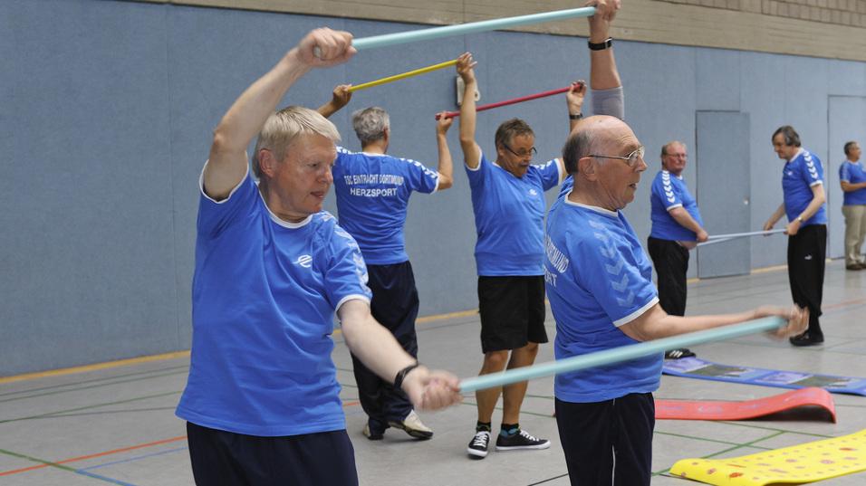 Ältere Männer in blauen T-Shirts trainieren mit Widerstandsbändern in einer Turnhalle.