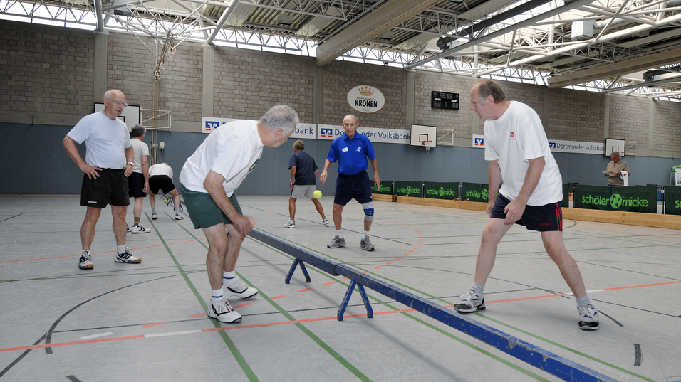 Gruppe &auml;lterer M&auml;nner trainiert auf einer Slackline in einer Sporthalle, w&auml;hrend einer den Ball wirft.