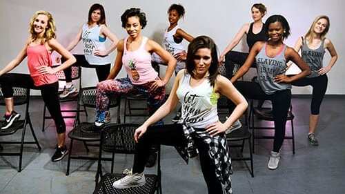 Gruppe junger Frauen in Sportkleidung posiert auf Stühlen in einem Tanzstudio. Farbiges Licht im Hintergrund.