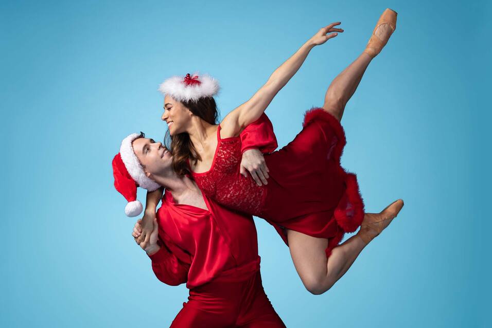 Ein Tänzer im Weihnachtsmann-Hut hebt eine Balletttänzerin in rotem Kleid mit Weihnachtsdekoration vor blauem Hintergrund.