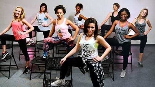 Gruppe junger Frauen in Sportkleidung posiert auf St&uuml;hlen in einem Tanzstudio. Farbiges Licht im Hintergrund.