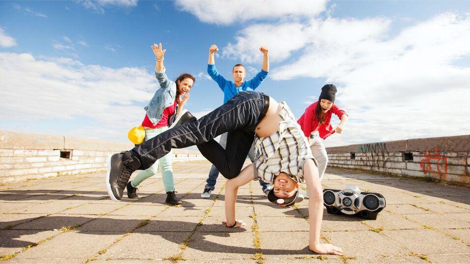 Vier junge Personen tanzen auf einer gepflasterten Fläche; einer macht einen Breakdance-Move, während die anderen applaudieren.