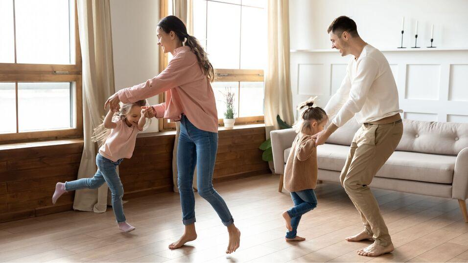 Familie tanzt gemeinsam im Wohnzimmer; zwei Kinder und Eltern lachen und halten sich an den H&auml;nden.