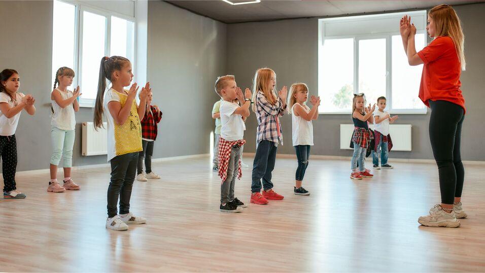Gruppe von Kindern in T-Shirts klatscht im Takt zu Musik in einem hellen Tanzraum.