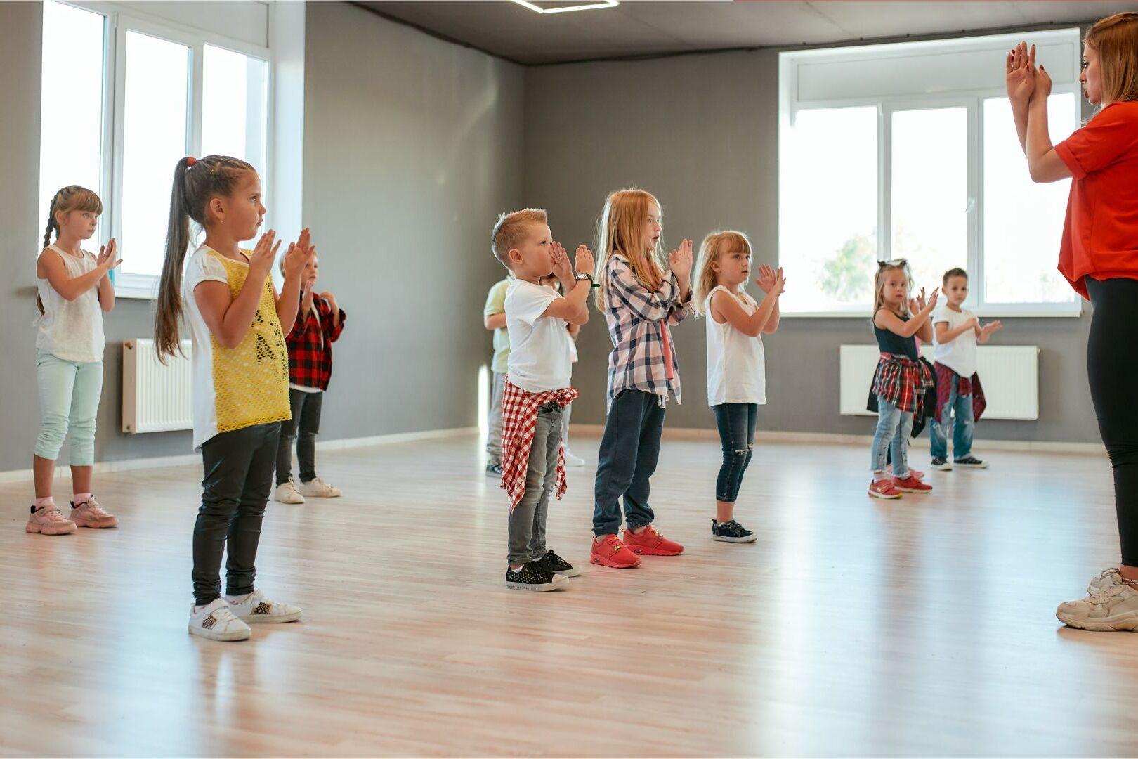 Gruppe von Kindern in T-Shirts klatscht im Takt zu Musik in einem hellen Tanzraum.