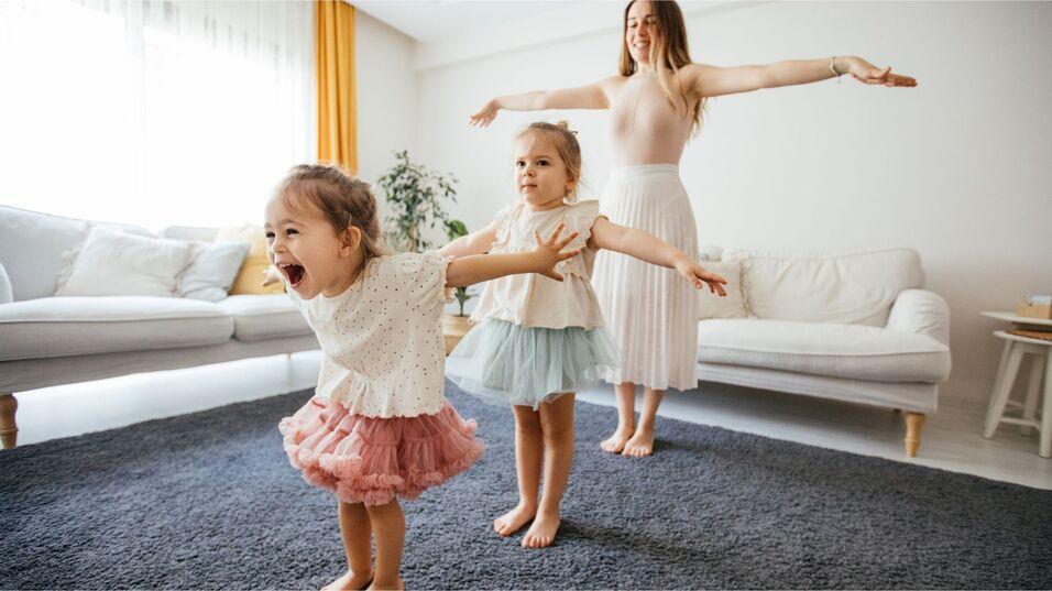Zwei M&auml;dchen in flauschigen Kleidern und eine Frau stehen in einem Wohnzimmer, sie machen spielerisch Bewegungen mit ausgebreiteten Armen.