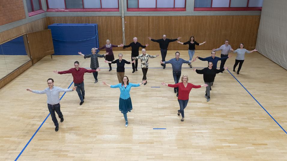 Eine Gruppe von 15 Menschen tanzt synchron in einem Sportraum mit Holzfußboden und großen Fenstern.