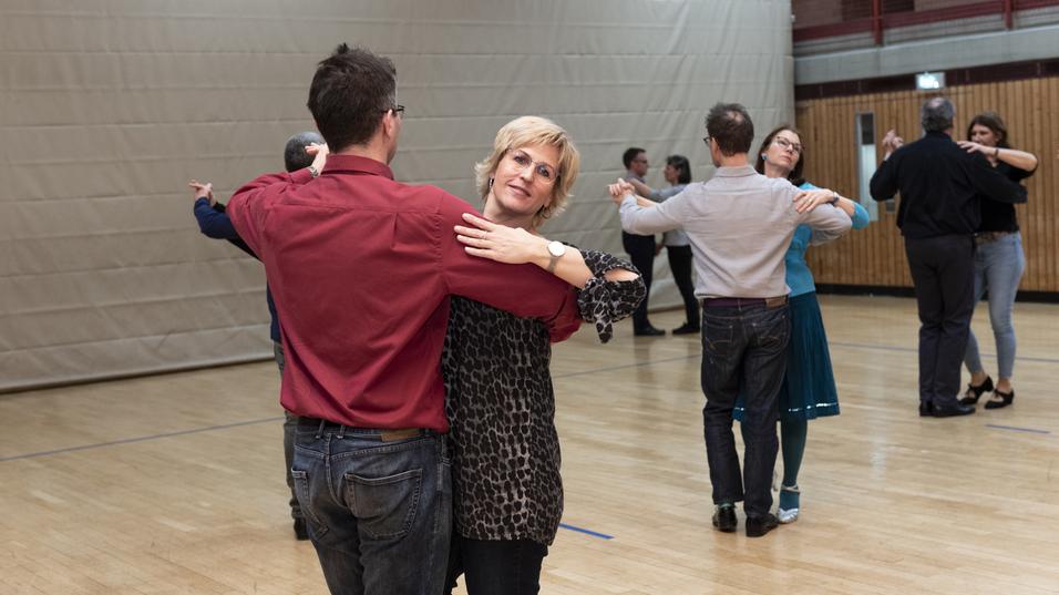 Tanzpaare üben im Gymnastikraum, eine Frau in Leo-Bluse tanzt mit einem Mann in rohem Hemd.