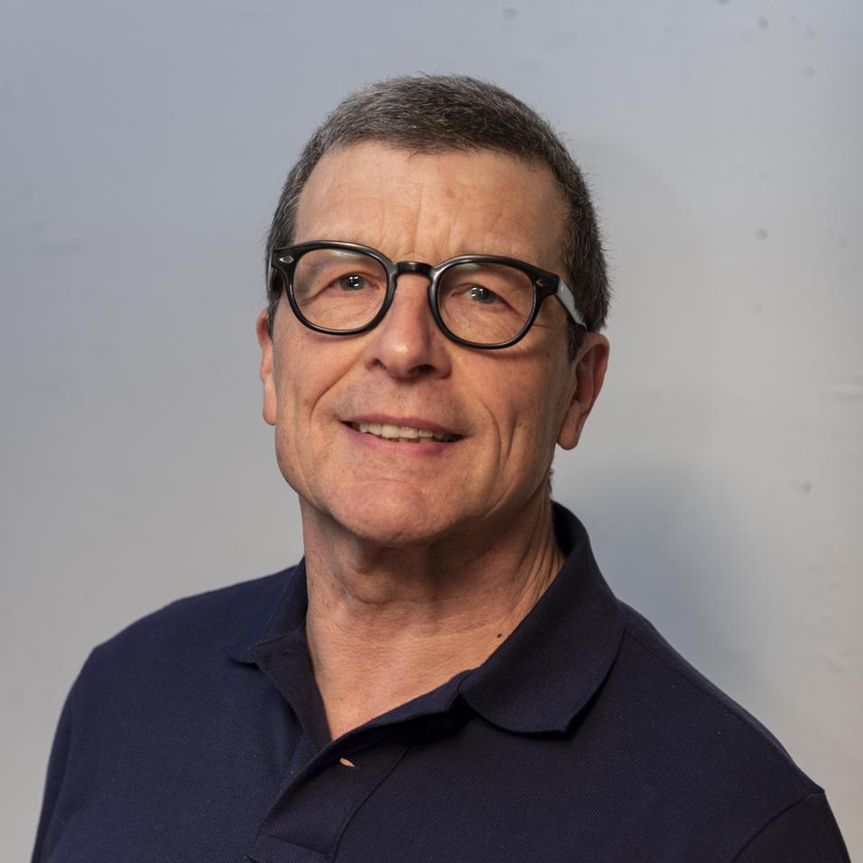 Portrait eines Mannes mit Brille, kurzen grauen Haaren und einem dunkelblauen Poloshirt vor einem hellen Hintergrund.